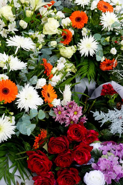 A vibrant floral arrangement featuring a mix of fresh flowers including bright orange gerbera daisies, white daisies, pale cream roses, pink chrysanthemums, deep red roses, and purple hydrangeas. The