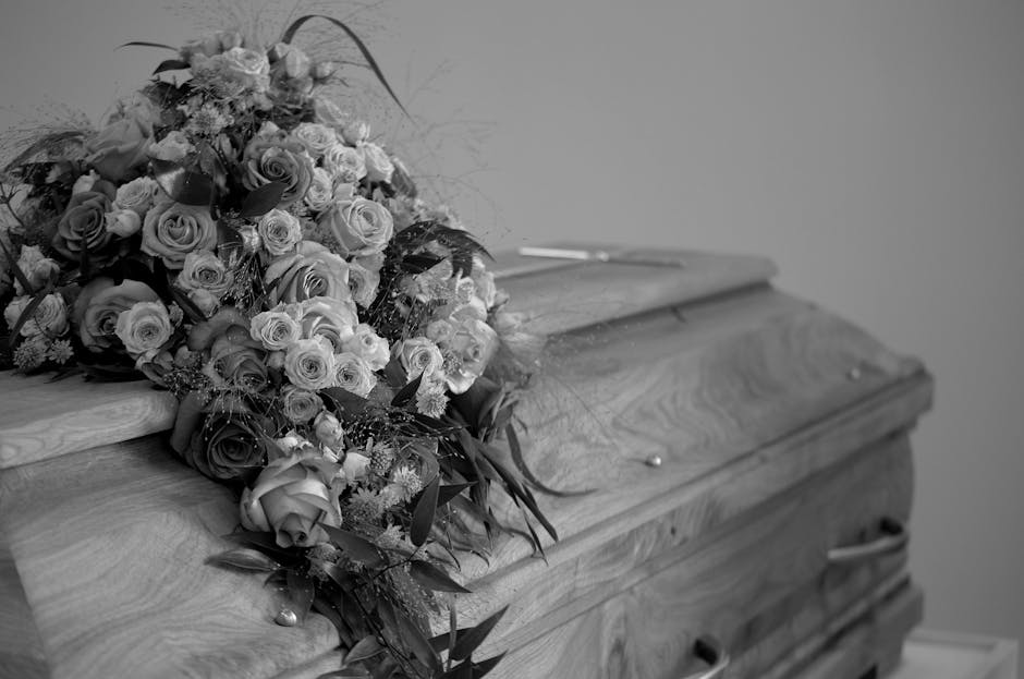 A large, elaborate floral arrangement resting on the lid of a wooden coffin. The bouquet features a variety of fresh flowers, predominantly roses, arranged in a cascading style. The roses are in shade