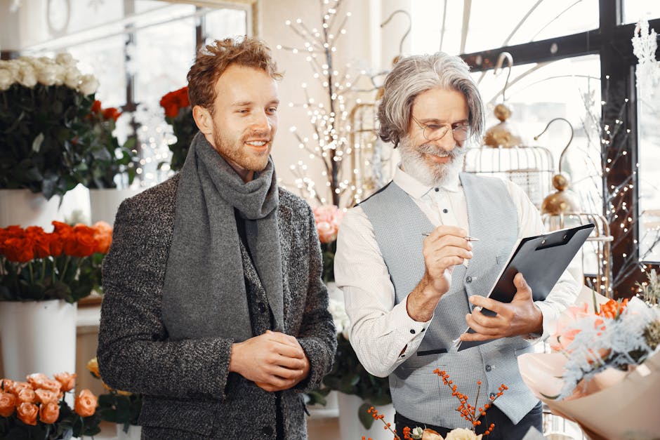 A male customer with short, light brown hair and a beard, dressed in a dark, textured coat and gray scarf, stands next to an older male florist with gray hair, glasses, and a beard, wearing a light bl