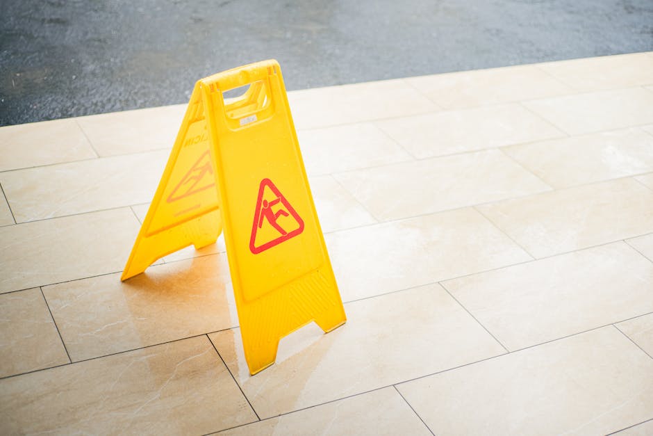 A yellow plastic wet floor caution sign with red triangular warning symbols stands on a light beige tiled floor, indicating a slippery surface. The sign is positioned in an indoor or outdoor environme