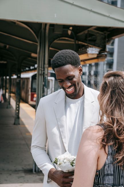 A young man dressed in a white suit is holding a small bouquet of white and green flowers, smiling warmly at a woman with long, wavy brown hair wearing a sleeveless plaid dress. They are standing outd