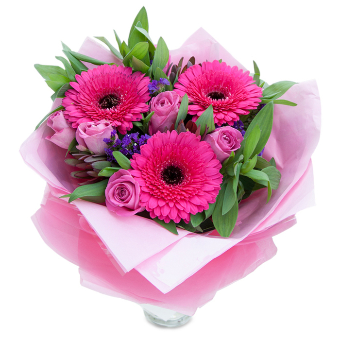 I often think that pink flowers can balance playfulness with a quiet grace, and this arrangement does both. Three fuchsia Gerbera daisies open wide, their many petals creating a starburst effect around deep, dark centres that anchor the eye. Nearby, roses in a muted lavender-pink tone curl inward with a softness that invites touch; their buds are fresh and poised between tightness and bloom. Sprigs of purple statice provide a delicate, textural counterpoint-tiny clustered blooms that nod like wildflowers tucked into a garden bouquet. The greenery feels intentionally garden-like: broad, glossy salal leaves give structure and a slightly waxy texture, while eucalyptus stems introduce a cooler, aromatic green that lifts the composition. The florist's wrap is visible-an inner layer of matte pale-pink tissue that cushions the stems, layered by a slightly crinkled, translucent fuchsia cellophane that adds a polished shimmer and echoes the flowers' hues. You can just see the top rim of a clear glass vase at the bottom of the image, so the bouquet reads as both hand-tied gift and ready-to-display centrepiece. The clean white background highlights the saturated pinks and purples and allows the shadows and curves of each petal to show. Ideal for placing on a coffee table in a home near Emerson Park Station or brightening a bedside in Upminster, this bouquet is the kind of thoughtful present people pick up before a visit to Hornchurch Country Park or a stop at Romford Market-a small, elegant way to celebrate someone you love.